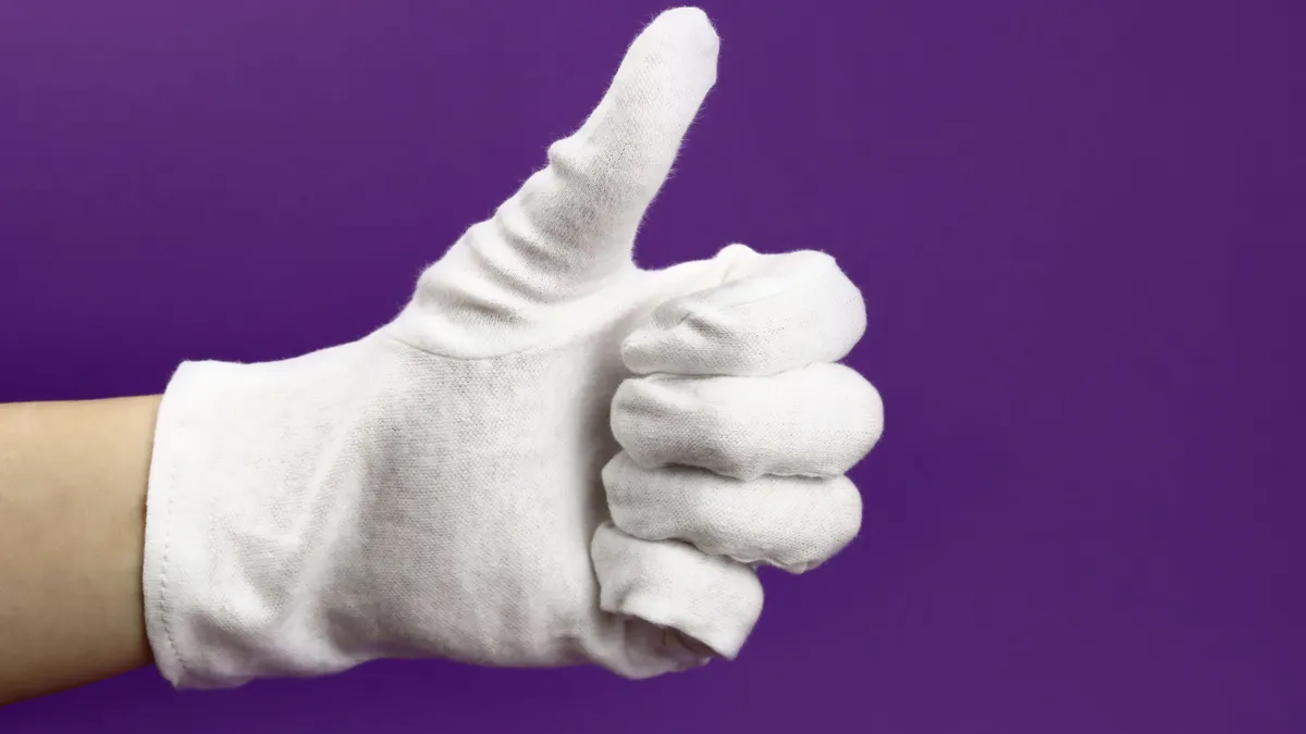 A hand wearing a white thumbs giving the thumbs up in front of a solid purple background