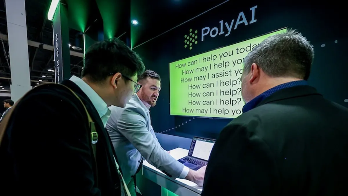 A man in a suite demonstrates a product called PolyAI at an exhibitor booth to other suited men.