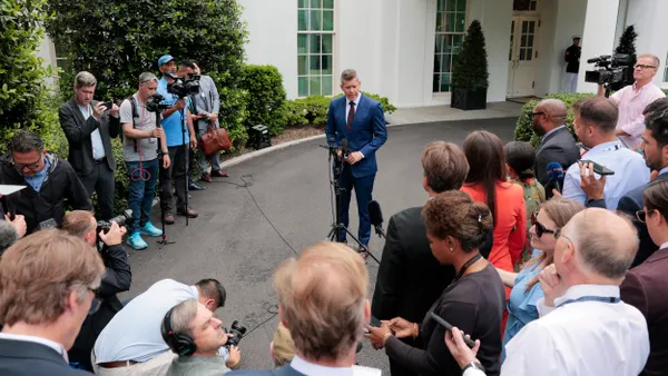Man in blue suit standing at a microphone surrounded by reporters with cameras outside the White House.