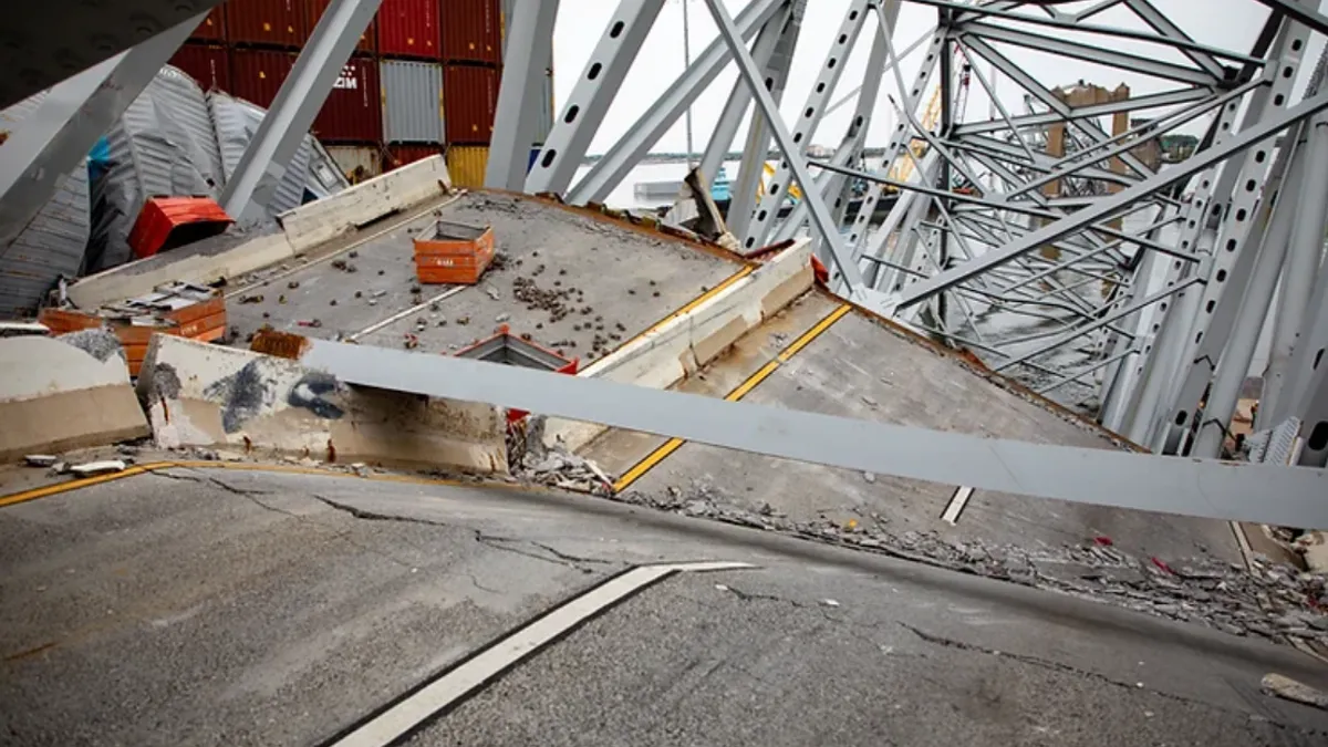 Street-level view shows buckled pavement and leaning bridge scaffolding, with cargo containers stacked in the background.