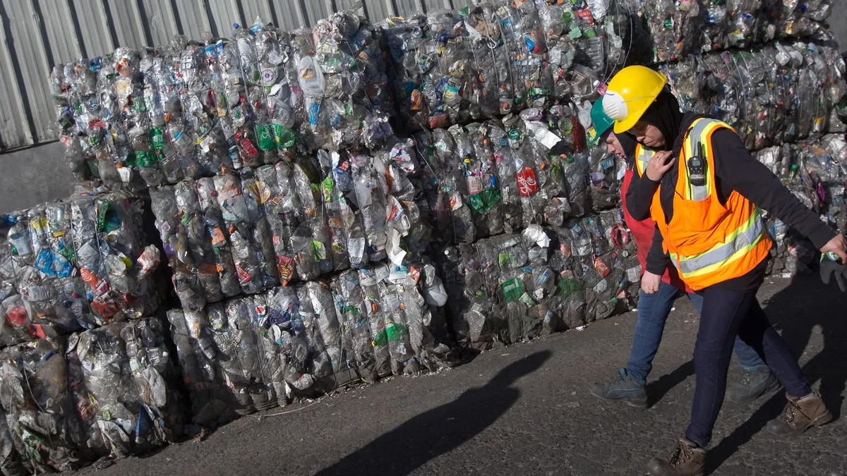 Two people with hard hats walking past bales of plastic scrap.