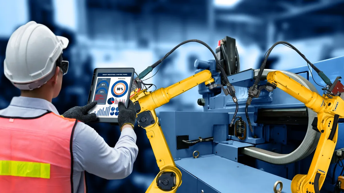 A person wearing a helmet, safety glasses, vest and gloves working on a portable device that powers the yellow robotic arms.