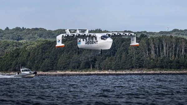 A white aircraft with 12 propellers labeled "Regent" flies above the water with a small boat in the distance and a tree-covered shoreline.