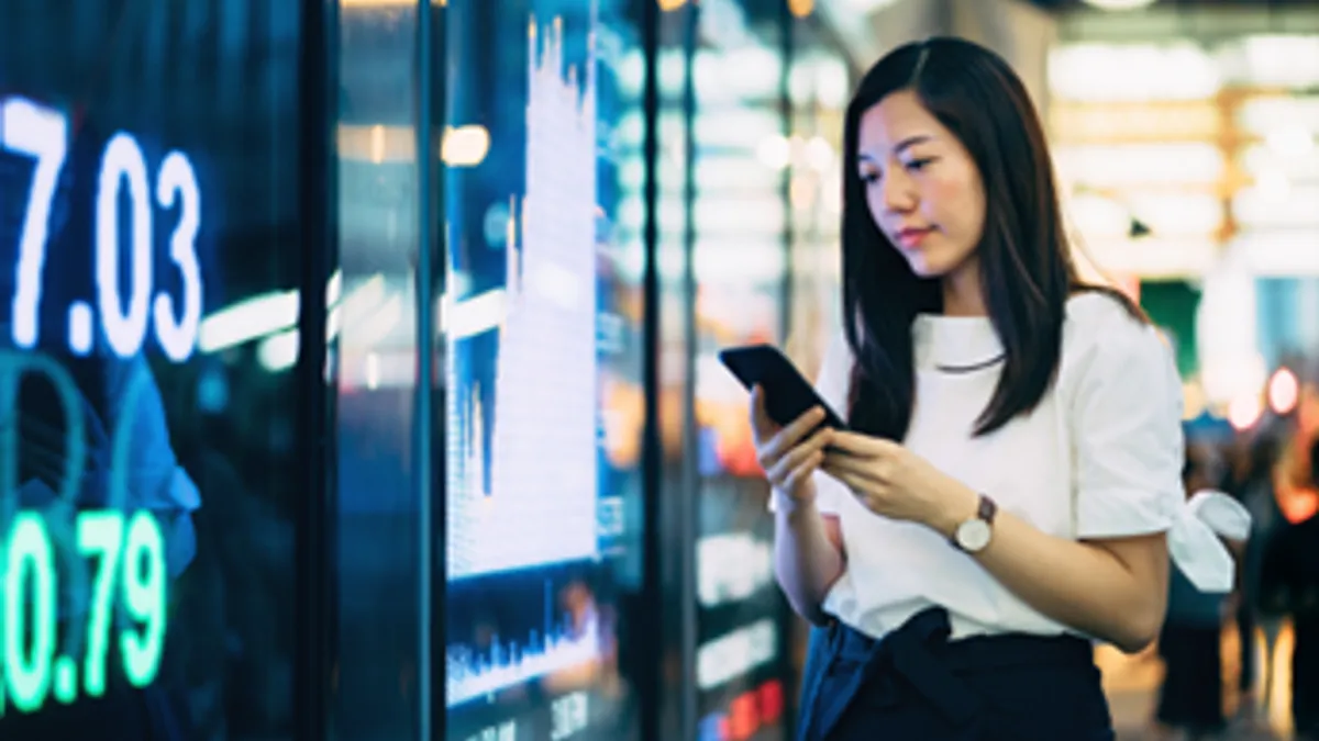 Confident business person checking financial trading data on smartphone by the stock exchange market display screen board in downtown financial district.