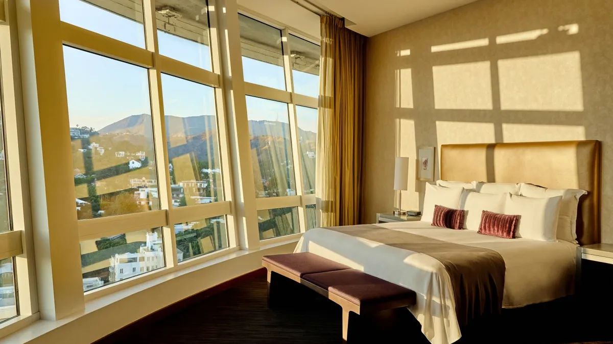 A hotel bed next to floor-to-ceiling windows overlooking Los Angeles.