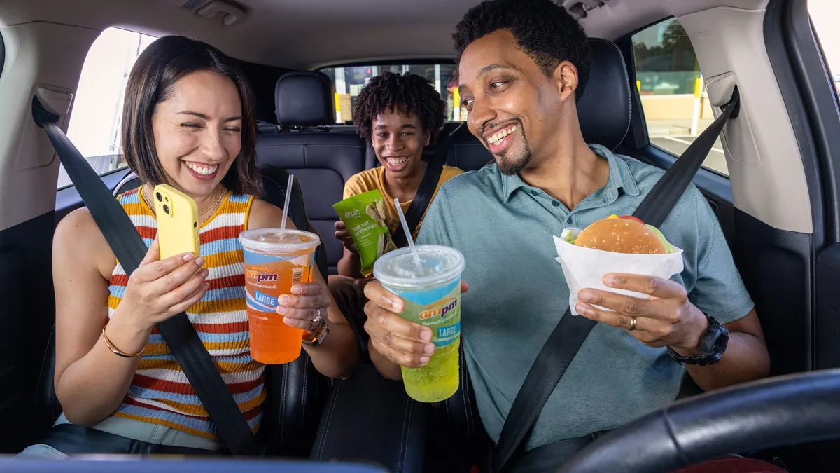 A photo of three people sitting in a car, smiling and holding food and drink items. The cups with drinks in them say ampm and Large, while the bag of nuts says epic sea salt pistachios.