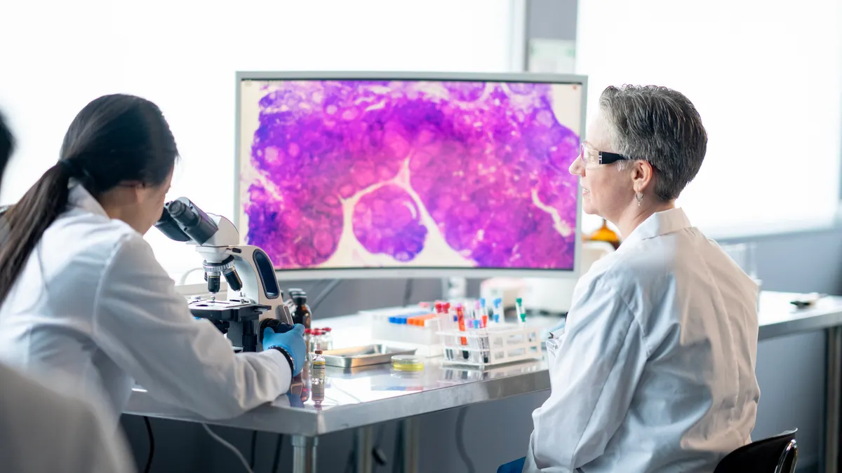 A person in a lab coat looks into a microscope while another person watches. A blown-up image of a cell is seen in the background on a computer monitor.