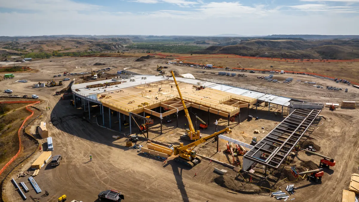 An overhead shot of a massive wooden building that stands on dirt in the middle of a bright day with clear skies.