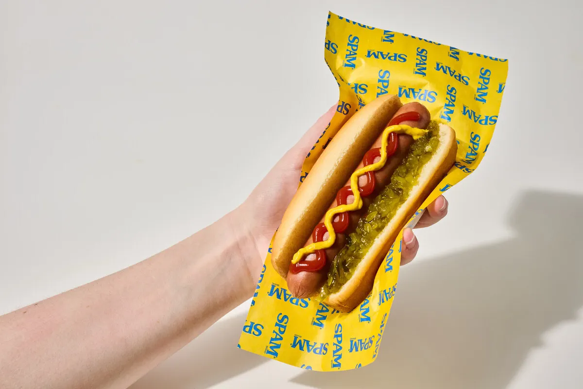 A person holding a Spam hot dog with ketchup, mustard and relish on a yellow wrapper