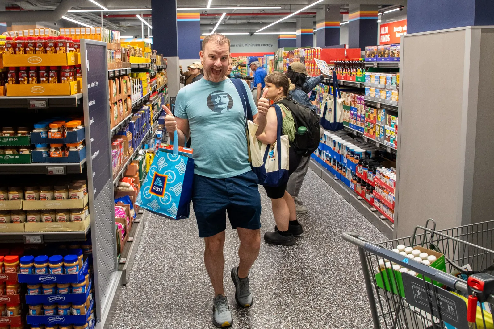 A very excited shopper in an Aldi aisle with an arm-full of bags giving the camera two thumbs up