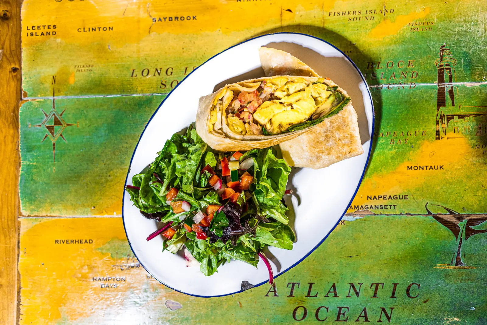 A plate on a table with a green and yellow map. A green salad is on the lower left side of the plate, and a cut-open burrito featuring scrambled Zero Egg is on the top right side.