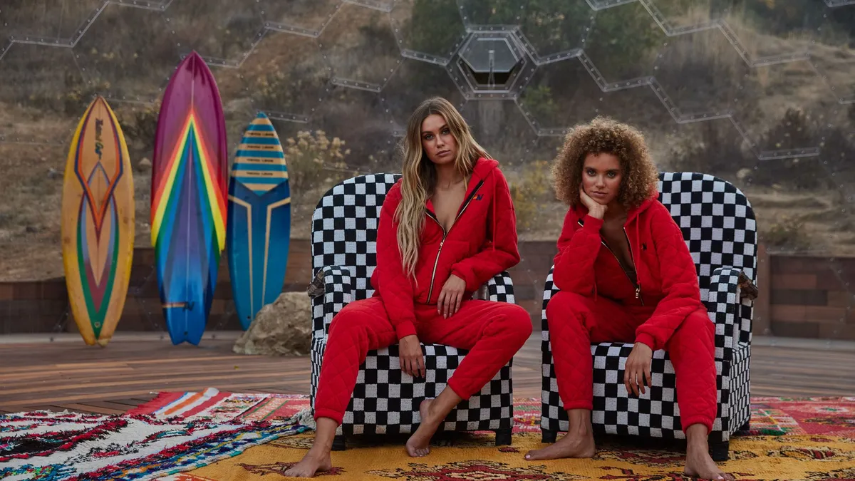 Two people are sitting in black and white checked chairs wearing red outfits. Three surfboards are on the left.