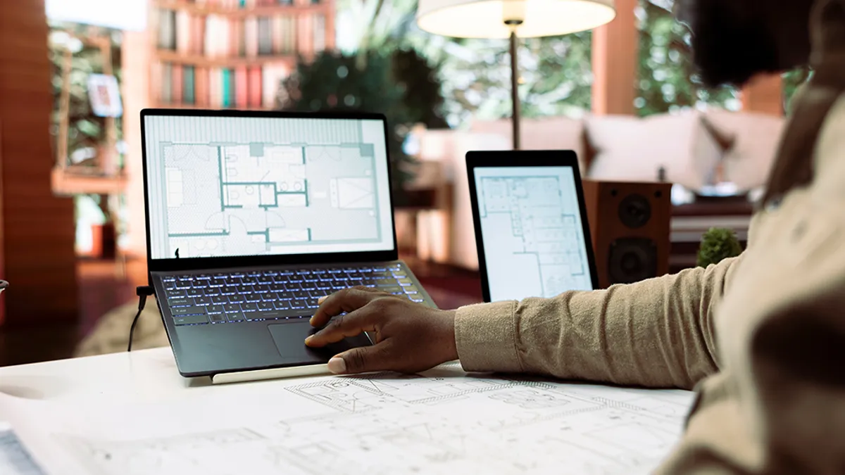 A person working on architectural plans displayed on a laptop and tablet, with blueprints laid out on the desk.