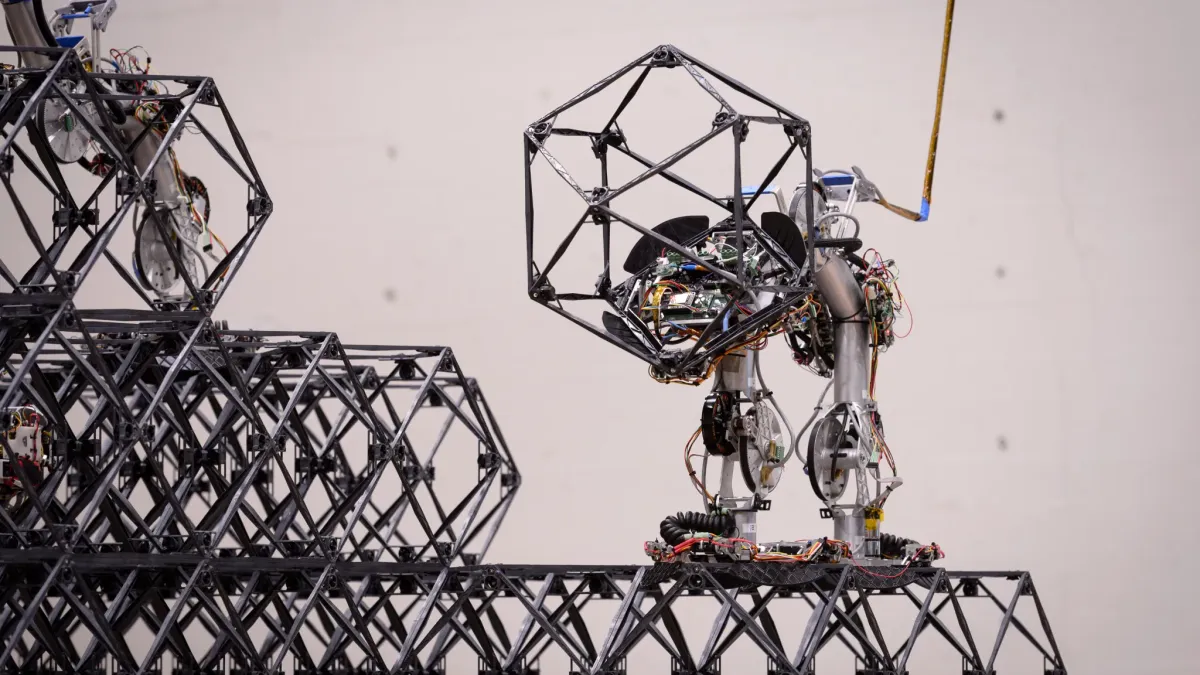 A robot, with plenty of exposed wires, sits atop a soccer ball-shaped wire frame and manipulates it indoors.