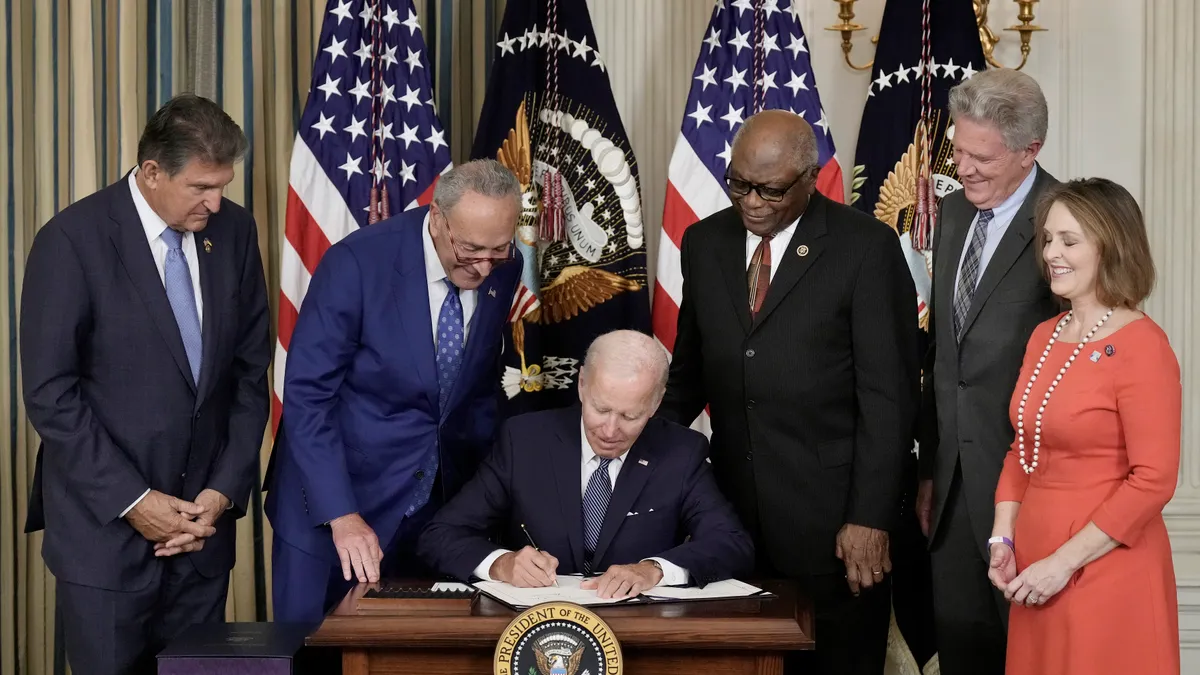 President Biden signs the Inflation Reduction Act surrounded by members of congress and backed by American flags in the White House.