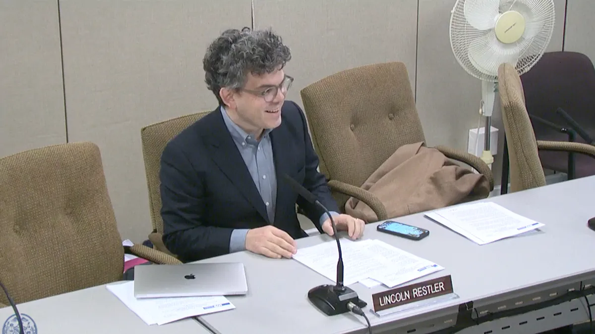 Lincoln Restler, member of the New York City Council, sits at a table in front of a microphone, speaking on a bill he introduced.