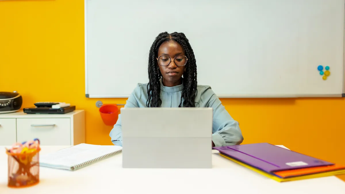 A teacher sits at a desk working on the computer in a classroom with a whiteboard in the background.