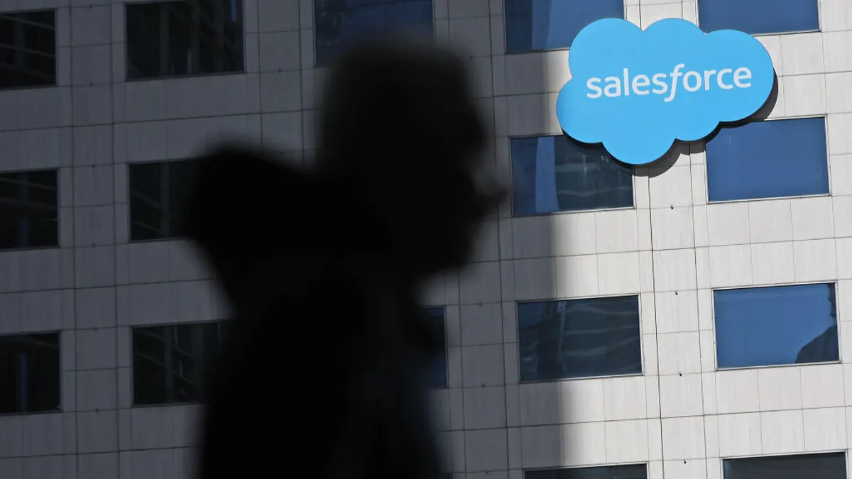 A corporate building bearing the logo of Salesforce in the background as the shadow of a person wearing a hoodie remains out of focus in the foreground