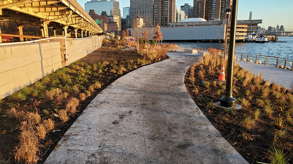 A grey footpath winds along the shore with a new york city skyline in the background.