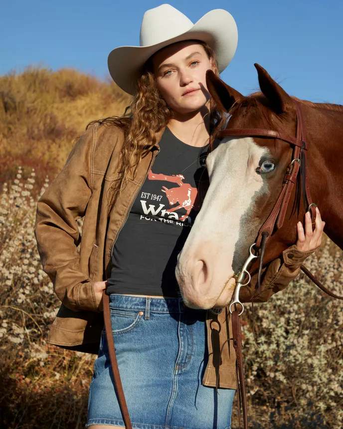 A model holds a horse&#x27;s head outdoors, wearing a cowboy hat, denim skirt, jacket and graphic T-shirt.