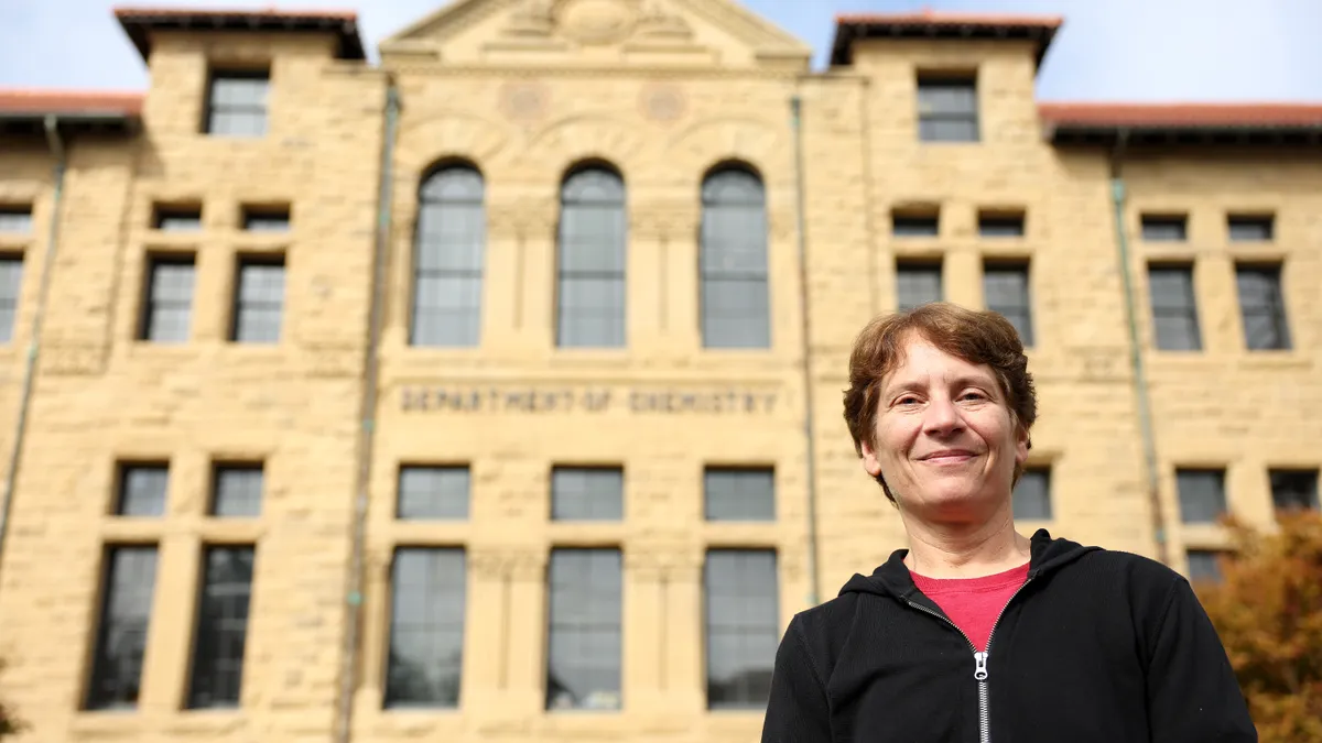 Wearing a black hoodie, Carolyn Bertozzi stands in the forefront with a tan brick building blurred behind.