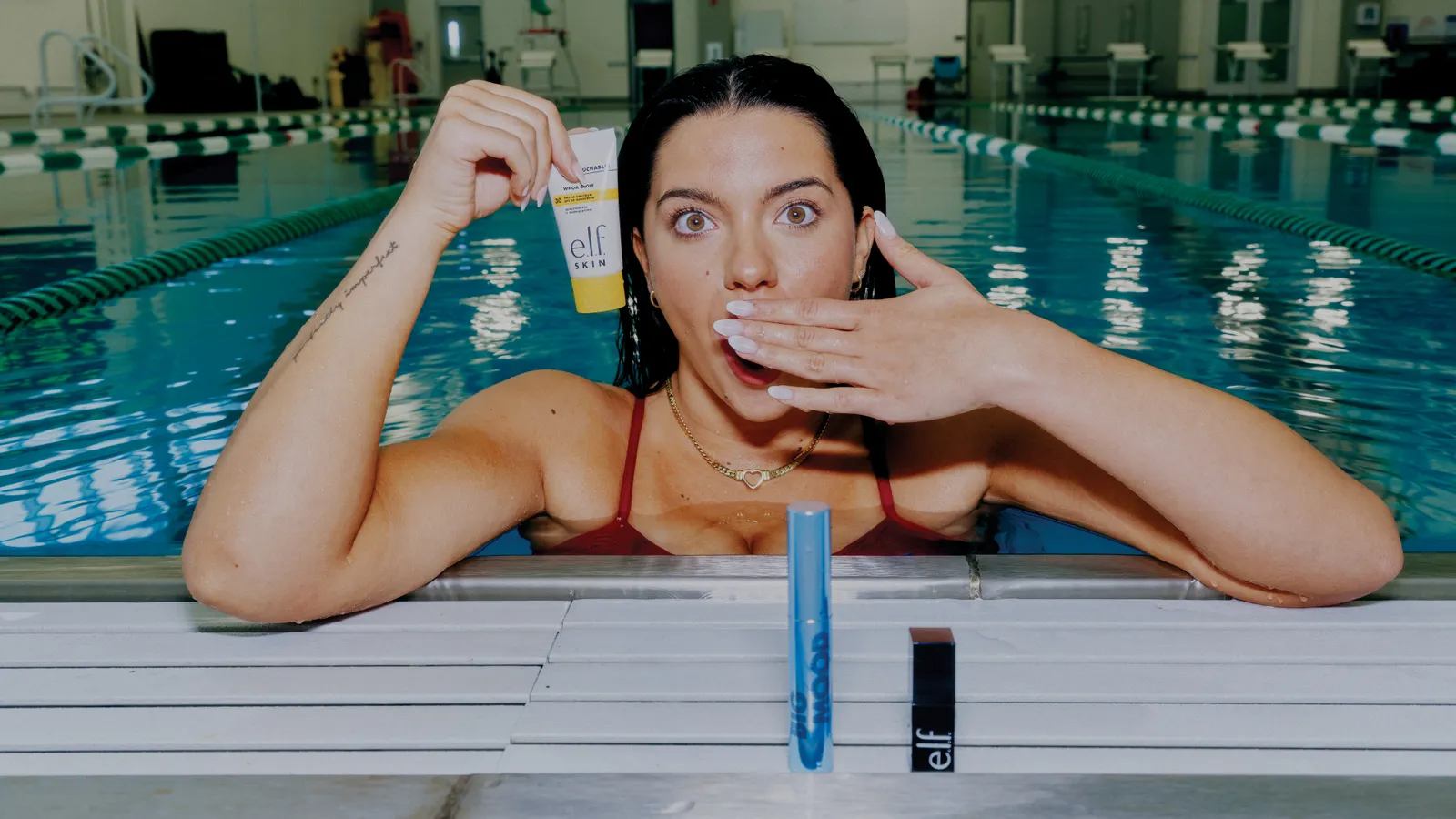 A person in a swimming pool holds a tube of e.l.f. sunscreen while a tube of mascara and a tube of lipstick sit on the pool deck.