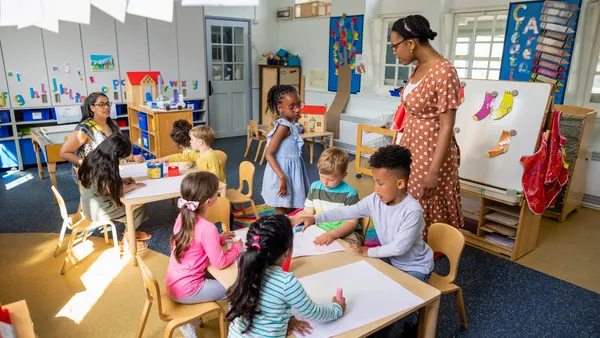 A teacher is standing in a classroom while several students and another teacher are seated at tables with papers on the tables.
