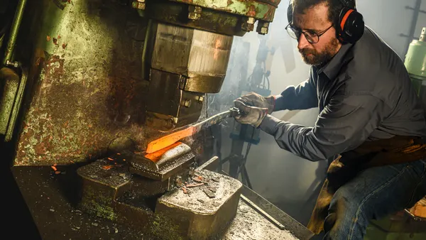 A person wearing ear protection, eyeglasses and gloves using a machine to shape heated steel.