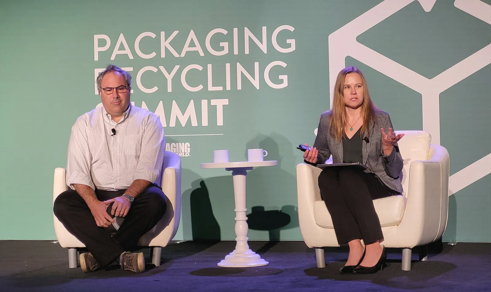 Two people sitting in chairs on a stage with a green background behind them that says "Packaging Recycling Summit."