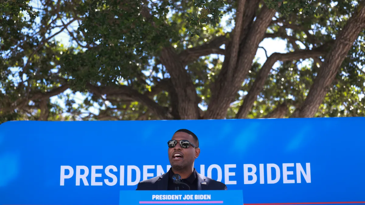 A man wearing sunglasses stands outside at a podium emblazoned with the words "President Joe Biden - Investing in America" in front of another Biden banner.