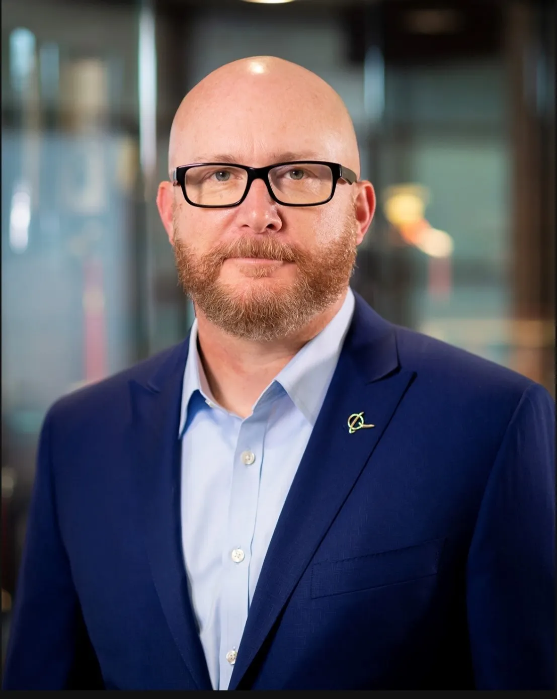 A headshot of a man with glasses and wearing a blue blazer.