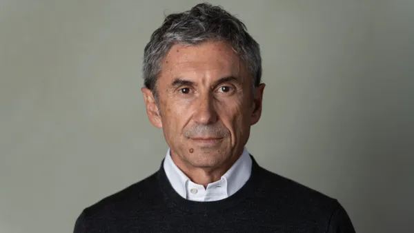 A person stares at the camera in a corporate headshot. He has short gray hair and wears a white shirt under a black sweater.