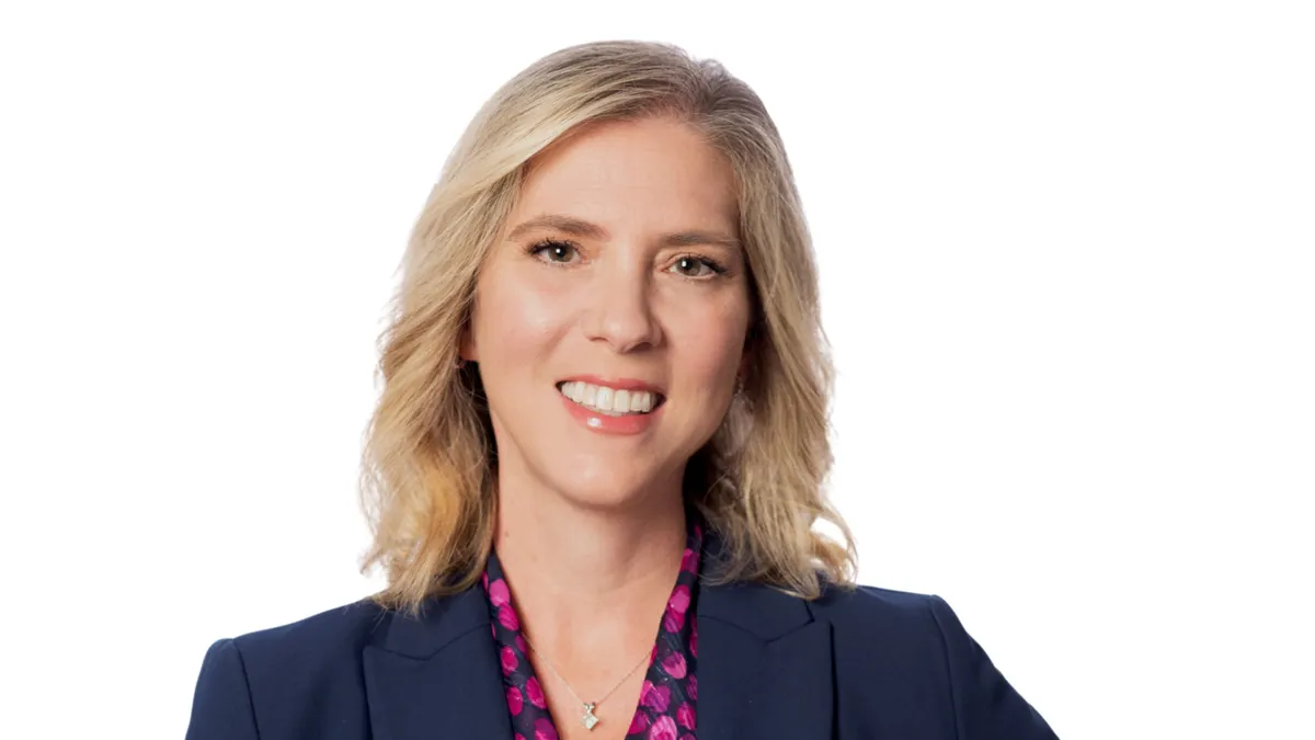 A person smiles while wearing a gray suit jacker and pink dotted blouse.
