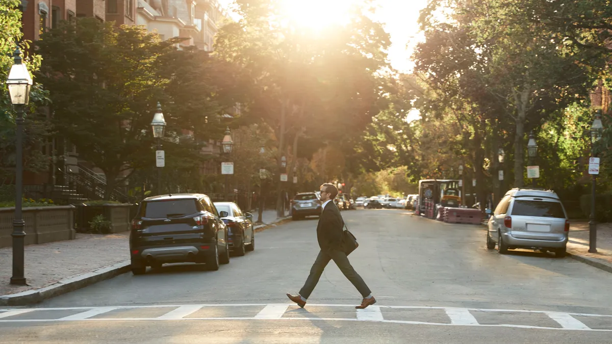 A person wearing business casual clothes and shoes walks across a sunlit road in a city