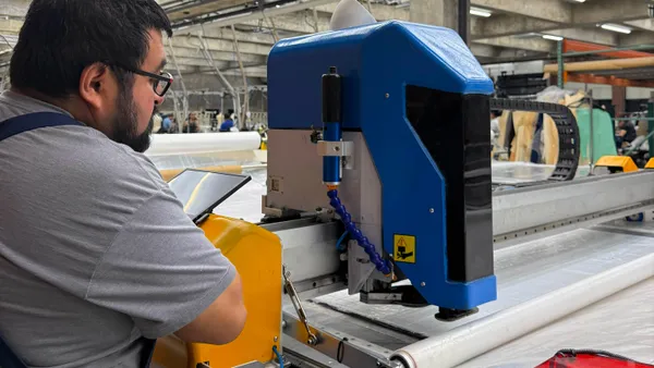 A worker uses an automatic cutter tool in an apparel factory setting. The equipment is a large blue apparatus that slides along a metal rod.