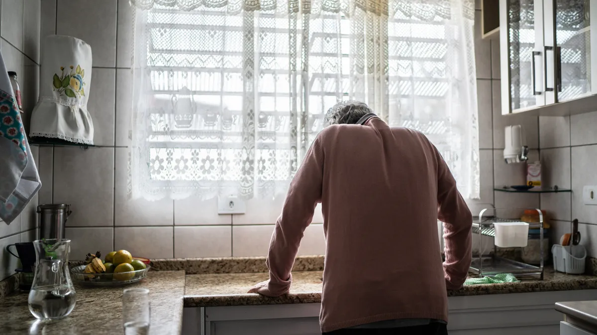 Older person in a kitchen facing toward a window with hands on the counter and head down