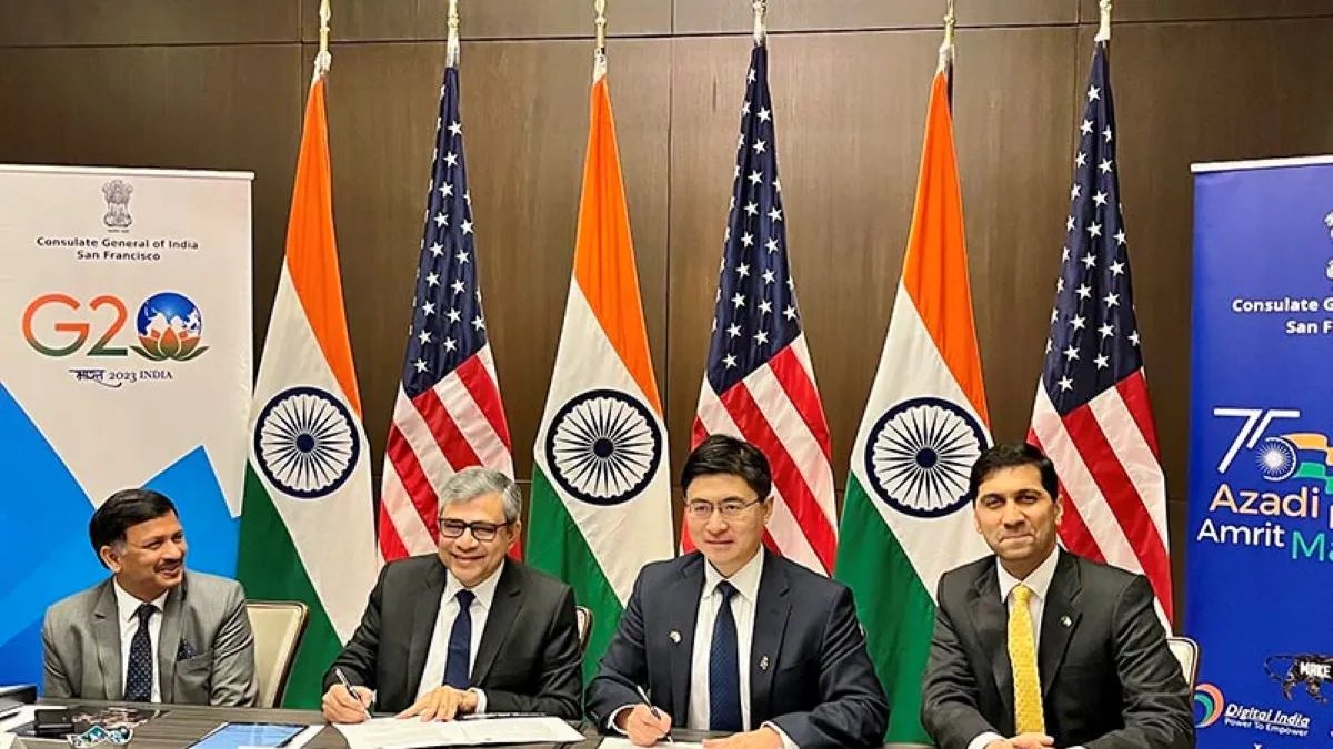 Four men in suits and tie at brown table signing documents, with the four U.S. and India flags behind them.