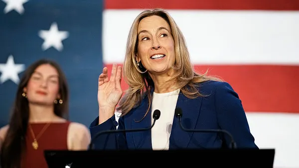 Blond woman in professional attire stands in front of a large American flag.