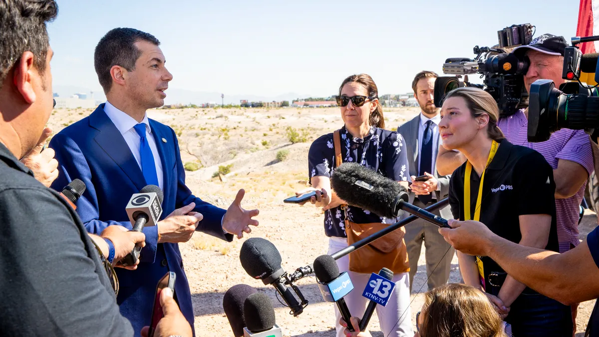 A man with brown hair and a blue suit speaks to a crowd of reporters.