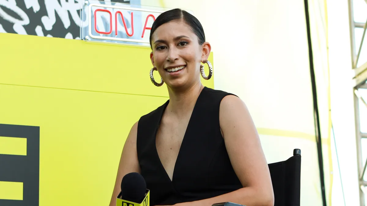 A woman with dark pulled back hair sits on a stage with a yellow backdrop smiling at the camera and holding a microphone.