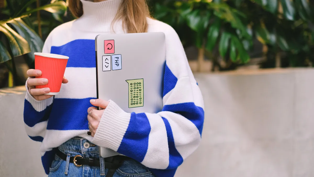 A faceless shot of a  woman in a striped long sleeve sweater holding a red and white coffee cup, and a closed laptop