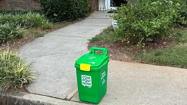 A small green bin with latched lid sits on the sidewalk near the curb outside a house.