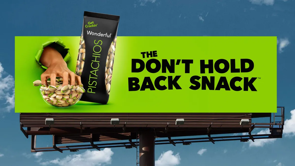 A billboard against a blue sky. Depicted on the green billboard is a hand reaching into a bowl of pistachios.