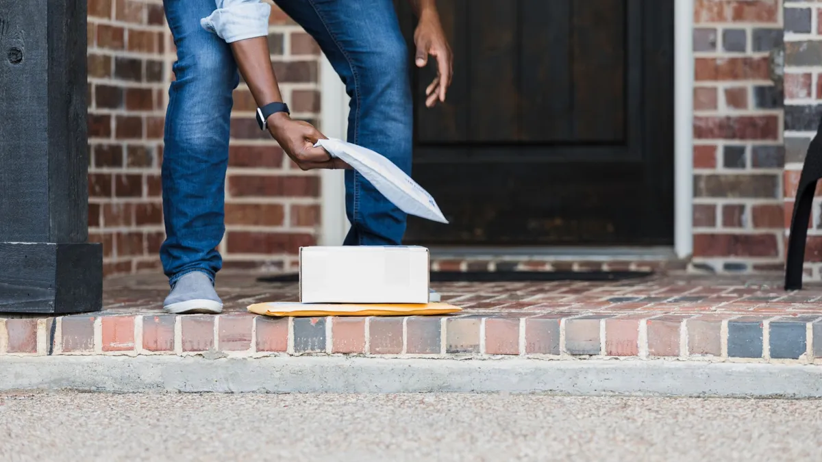 person bends over and picks up a stack of packages from a porch step