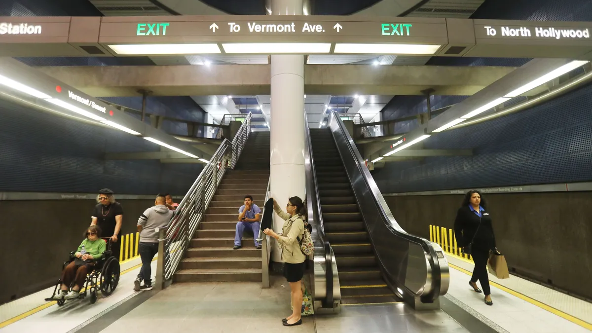 A sparse amount of people linger on an underground platform, reachable by stairs, next to two sets of train tracks.