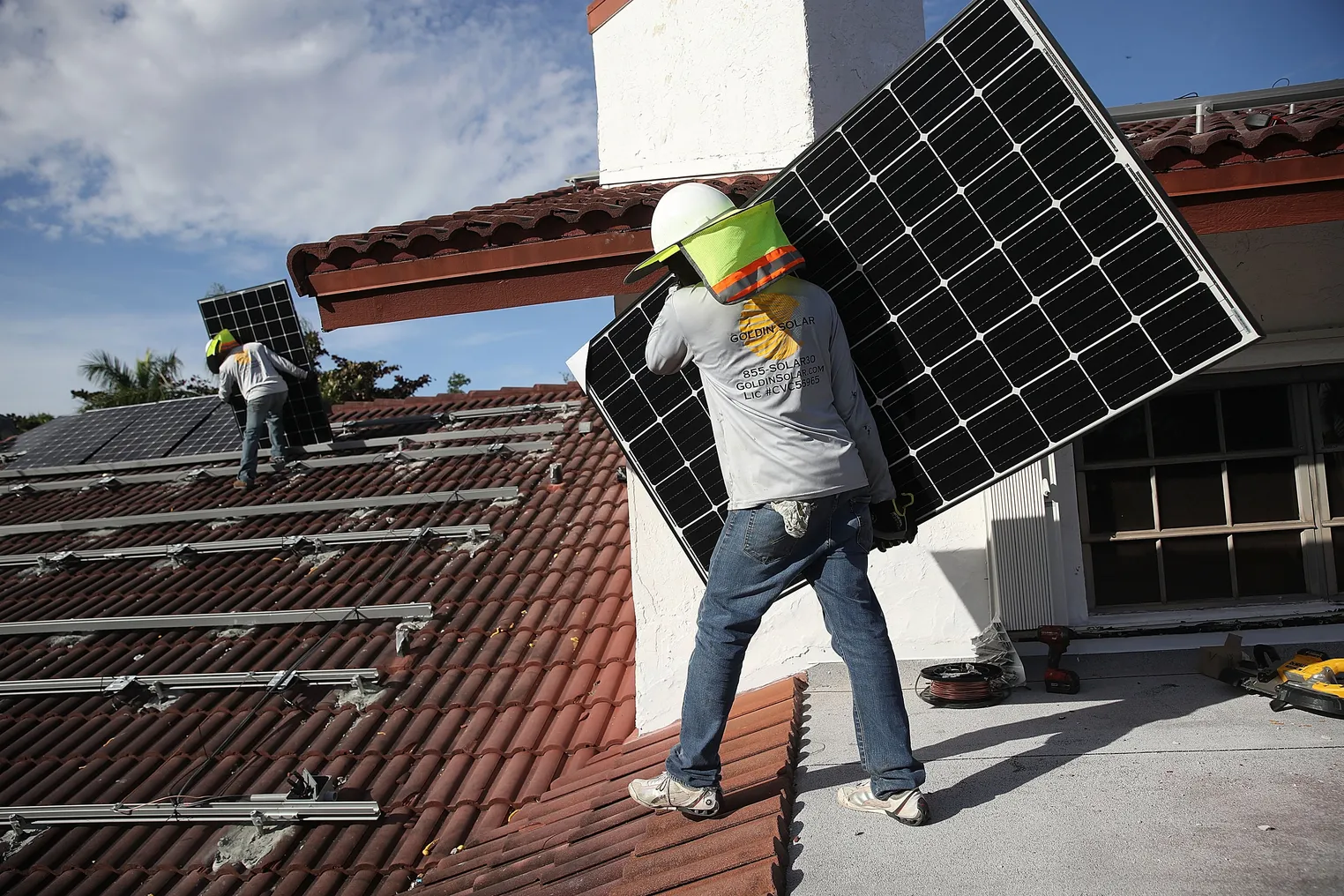 Two workers standing on a roof holding solar panels for installation in a metal framework on the roof.