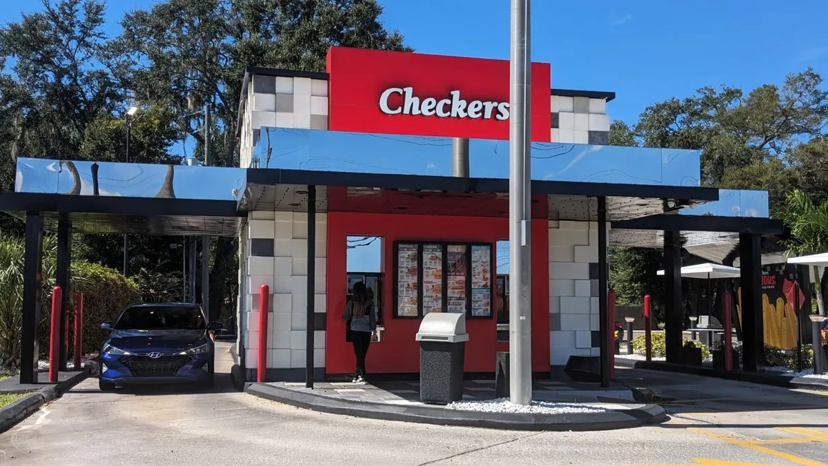 An image of a small restaurant with a red facade that has a white logo saying Checkers. There is a woman waiting at a pickup window and a blue car at a drive-thru window.