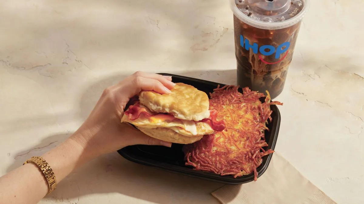 A woman's hand is holding a breakfast biscuit in a black takeout container with hashbrowns from IHOP.