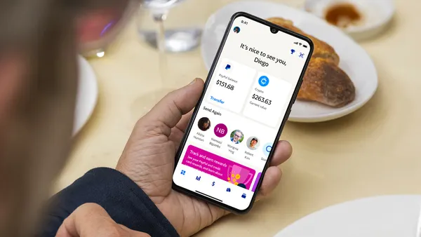 A hand holds a phone on a kitchen table. The screen reads, "It's nice to see you, Diego! PayPal balance: $151.68, Crypto: $263.63. Send again."