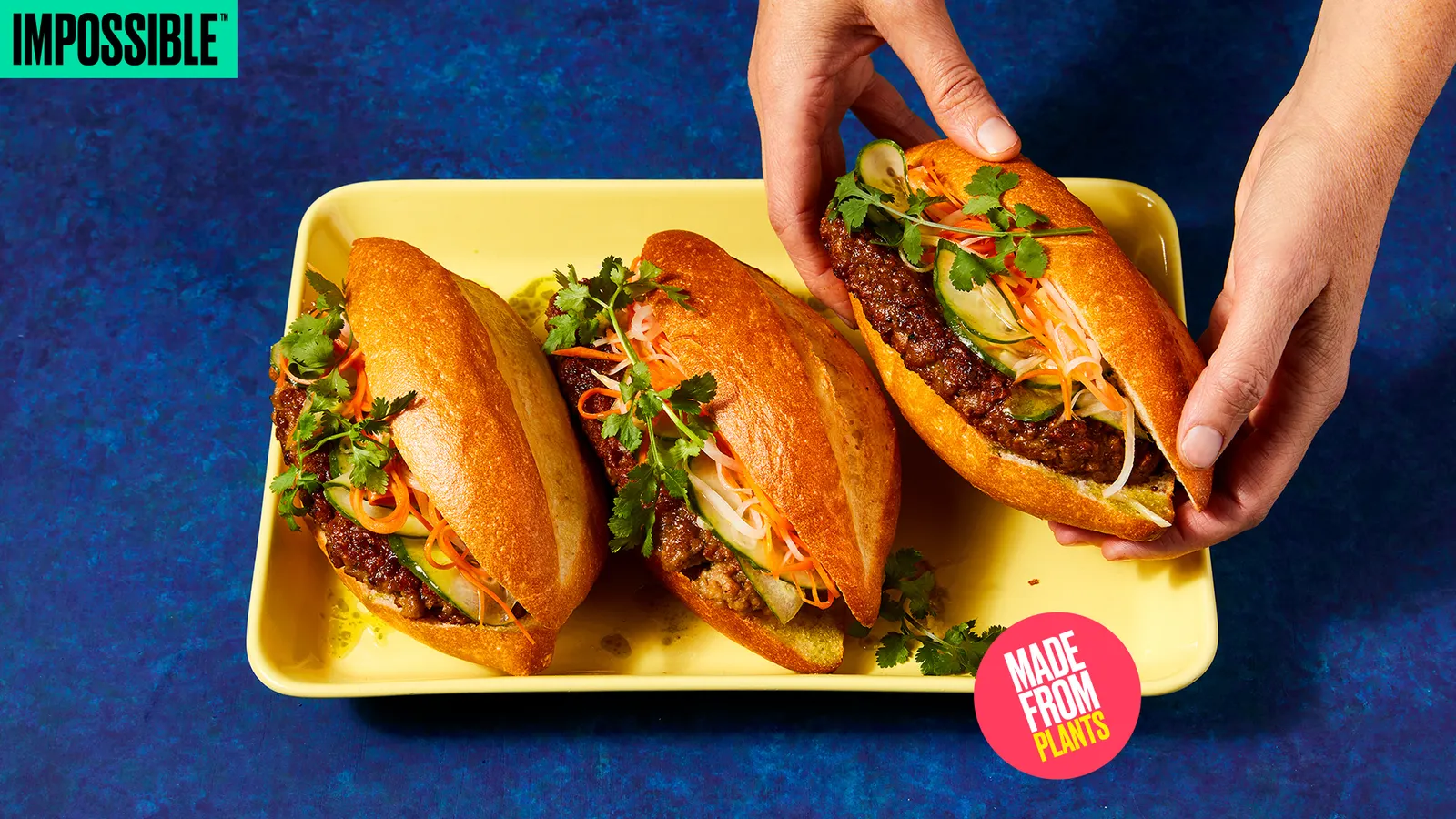 A hand picks up one of three bahn mi sandwiches made with Impossible Pork. All three sandwiches are on a yellow green rectangular plate on a royal blue table.
